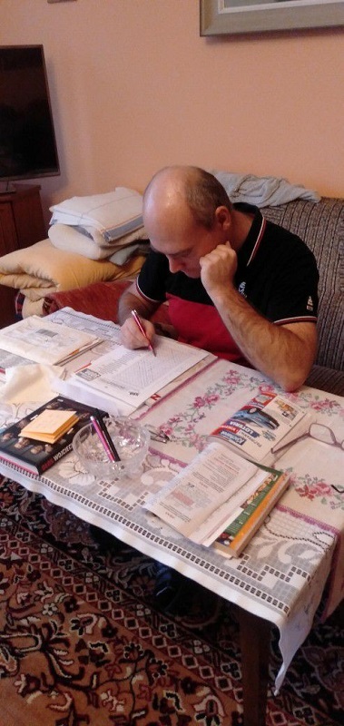 Mężczyzna siedzący na kanapie rozwiązuje pisemne zadania, na stole leżą materiały edukacyjne.