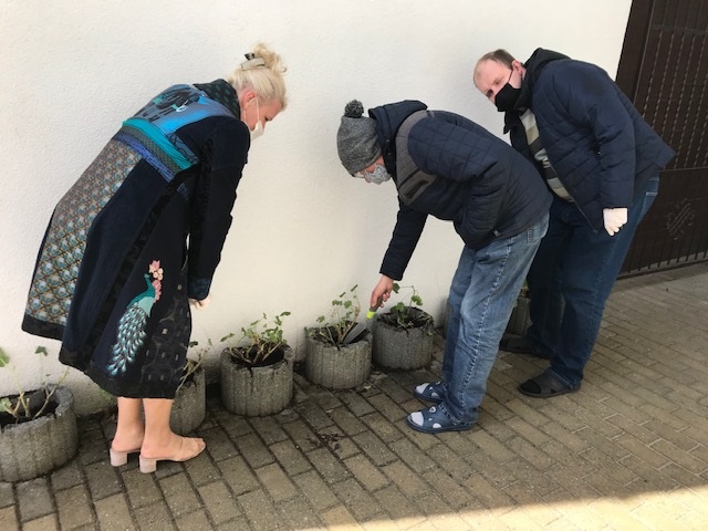 Dwóch mężczyzn i jedna kobieta sadzą kwiaty ogrodowe do donic betonowych przed wejściem do ośrodka.