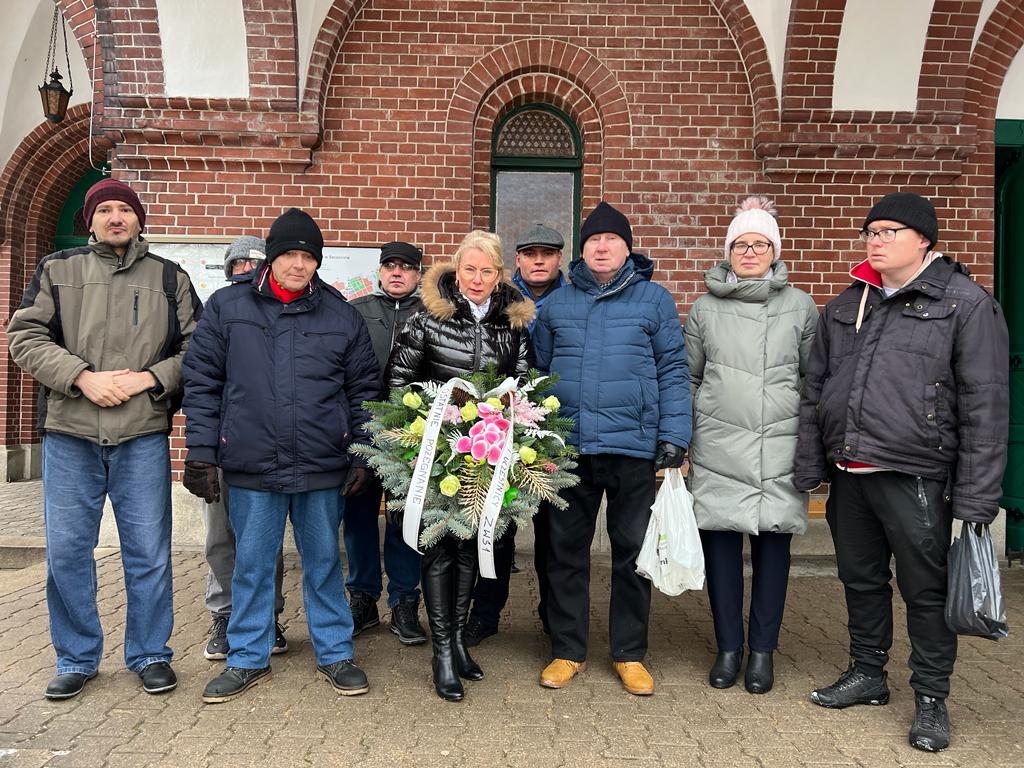 Grupa kobiet i mężczyzn w zimowych kurtkach i czapkach stoją przed kaplicą. Po środku stoi kobieta z wieńcem pogrzebowym. 