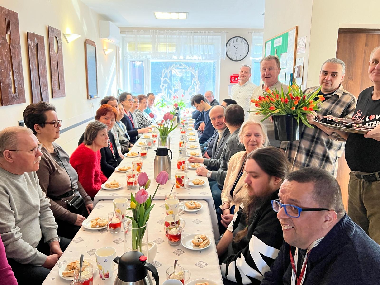 grupa kobiet i mężczyzn siedzą przy zastawionym stole. Po prawej panowie rozdają tulipany kobietom. 