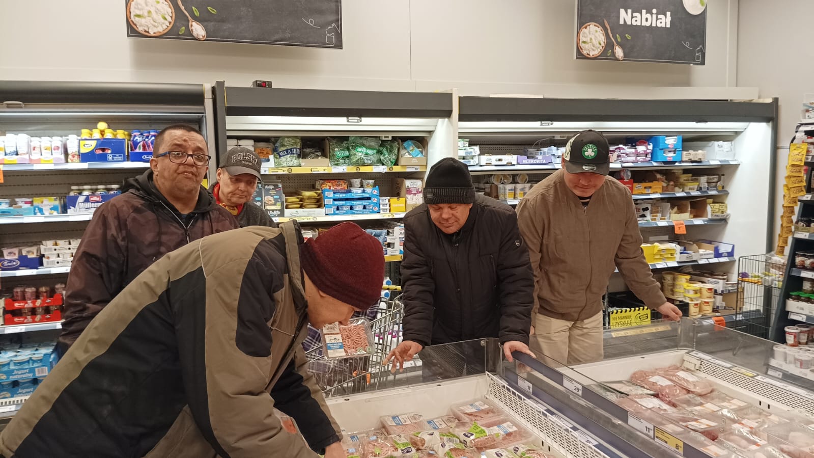 Pięciu mężczyzn w sklepie na dziale z nabiałem oglądają produkty w lodówkach.