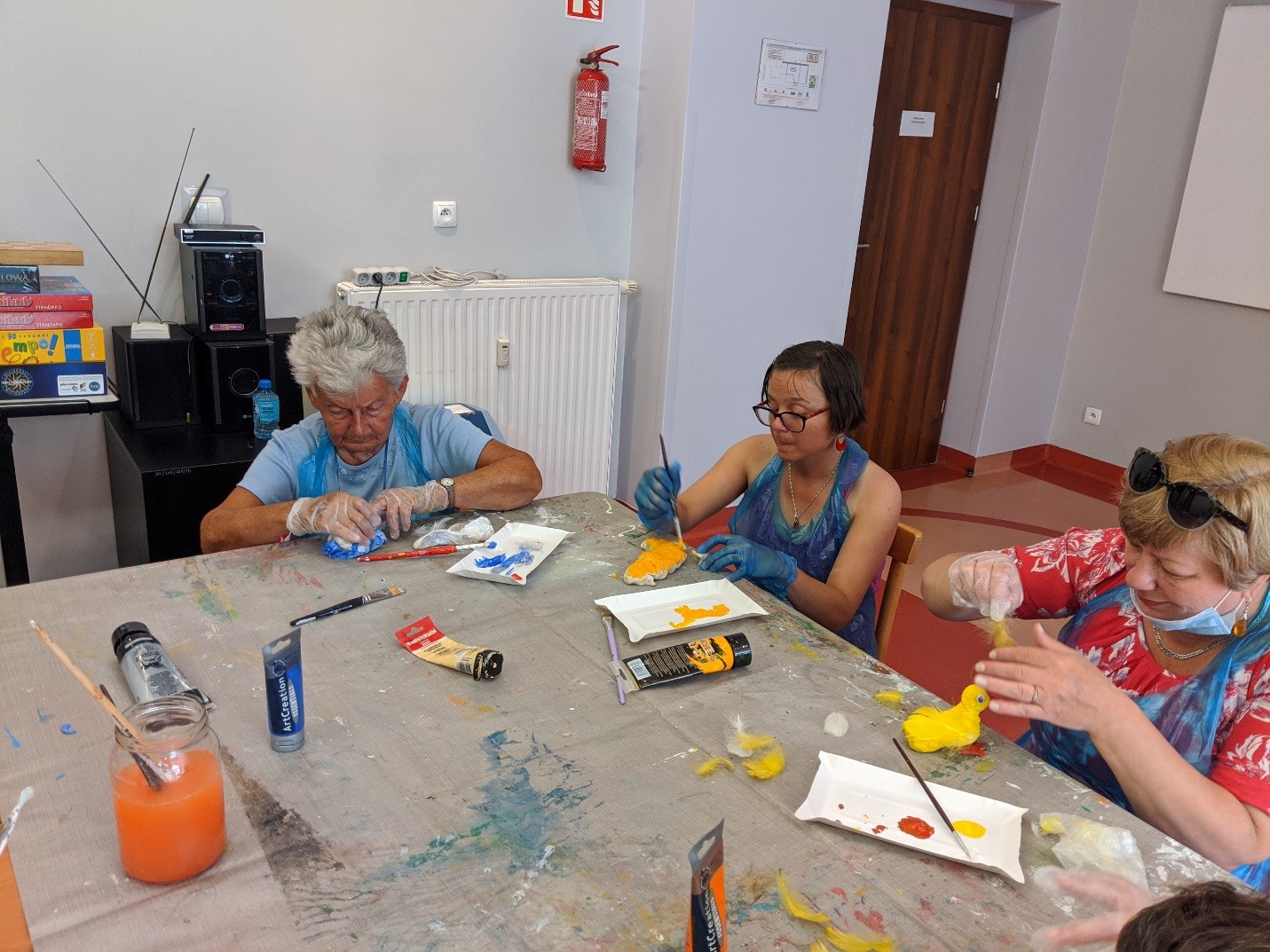 Trzy kobiety siedzące przy stole podczas warsztatów plastycznych malują farbami. 