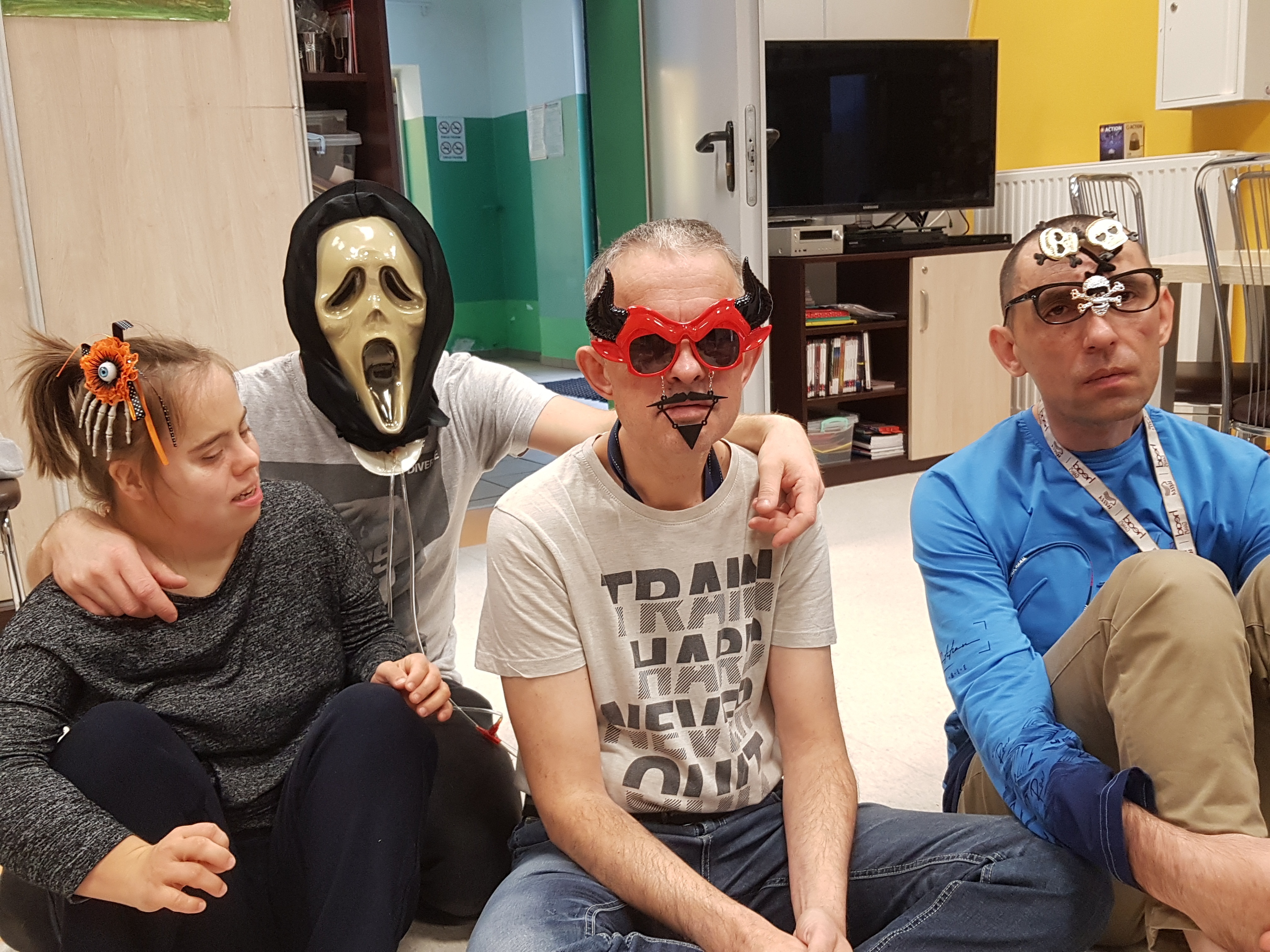 W sali siedzi troje uczestników pomiędzy którymi kuca terapeuta. Od lewej strony, pierwsza osoba ma na głowie opaskę, druga maskę, trzecia maskę, a czwarta ozdobne okulary.