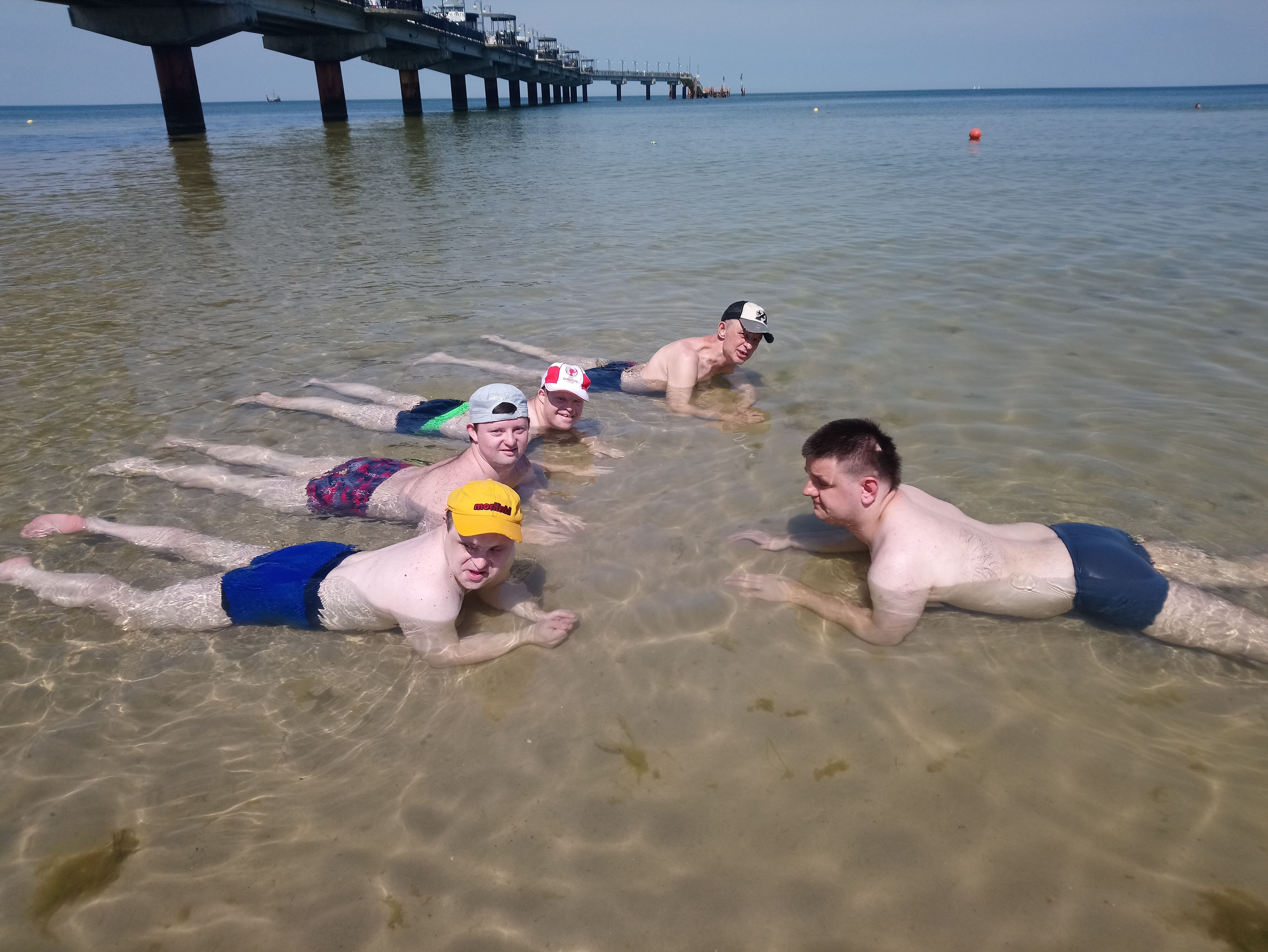 5 uczestników leży na brzuchach na brzegu morza. Ubrani są w kąpielówki, a na głowach mają czapki. 