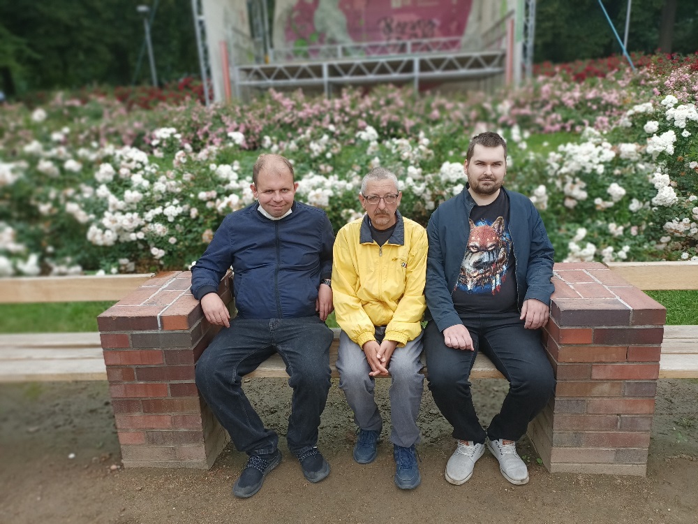 Trzej mężczyźni siedzą na ławce na tle róż.