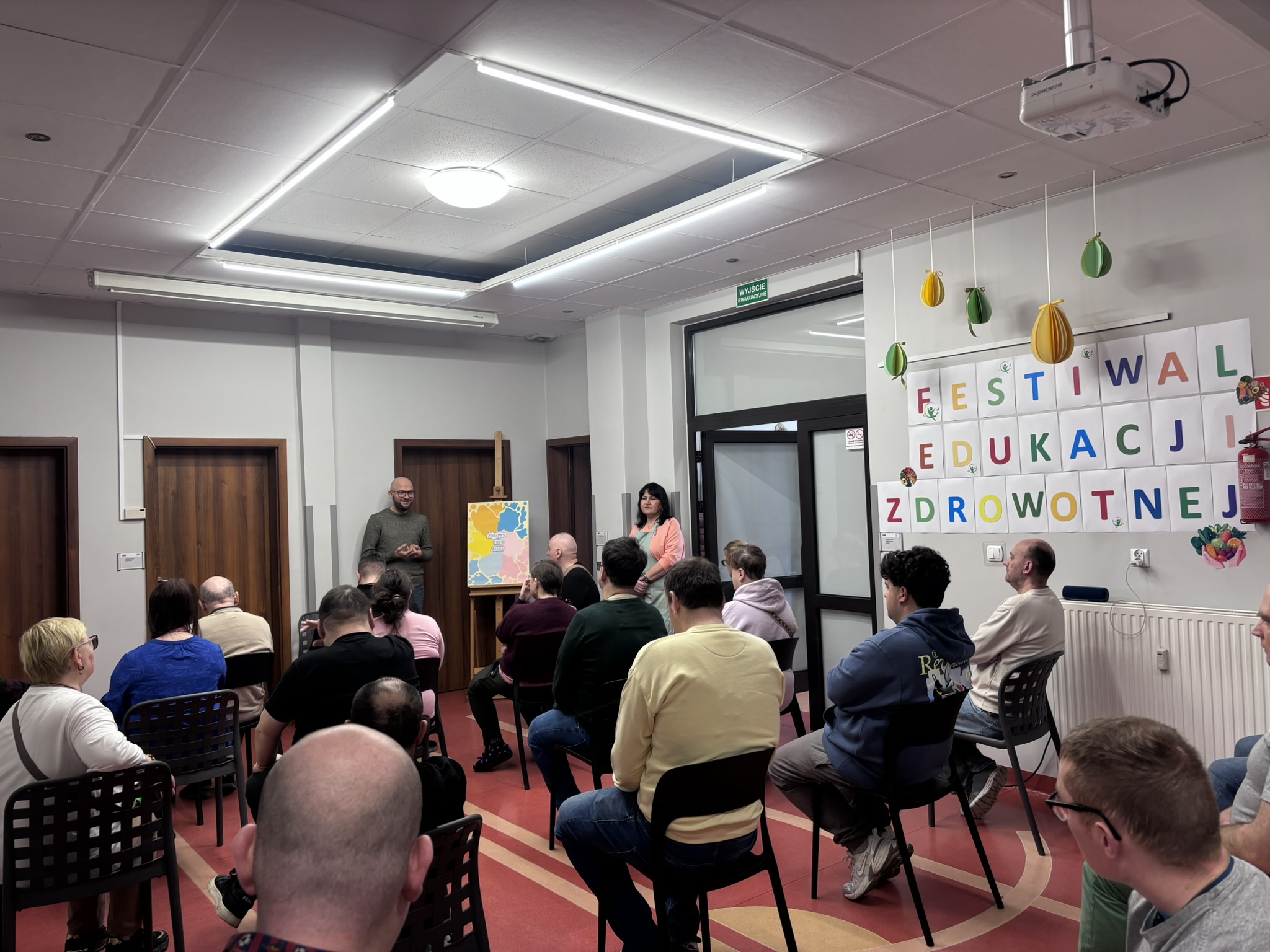 Na zdjęciu widać grupę ludzi. Ludzie na pierwszym planie siedzą na krzesłach. W tle widać dwie stojące osoby. Na ścianie widnieje kolorowy napis: FESTIWAL EDUKACJI ZDROWOTNEJ.
