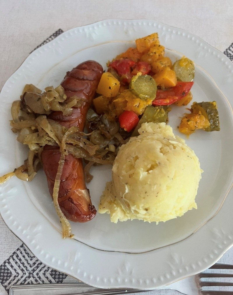 Na białym talerzu znajduje się porcja purée ziemniaczanego, grillowana kiełbasa z podsmażoną cebulą oraz kolorowa mieszanka warzyw: papryki, ogórka i dyni.