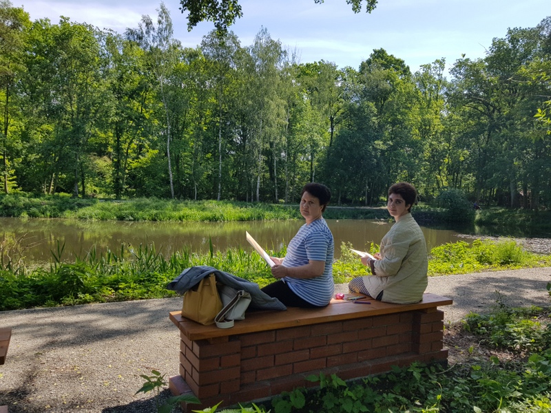 Dwie kobiety siedzące nad jeziorem w otoczeniu zieleni, w ręku trzymają kartki.