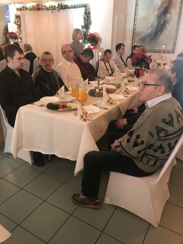 Kobiety i mężczyźni siedzący za świątecznym stołem, na którym znajdują się bożonarodzeniowe potrawy. 