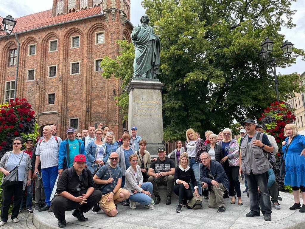 Grupa kobiet i mężczyzn pozujących do zdjęcia pod pomnikiem Mikołaja Kopernika w Toruniu. 