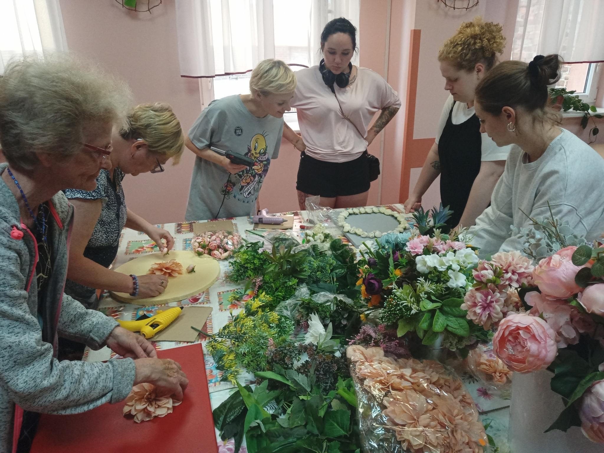 Kobiety stojące wokół stołu, tworzące kompozycje ze sztucznych kwiatów.