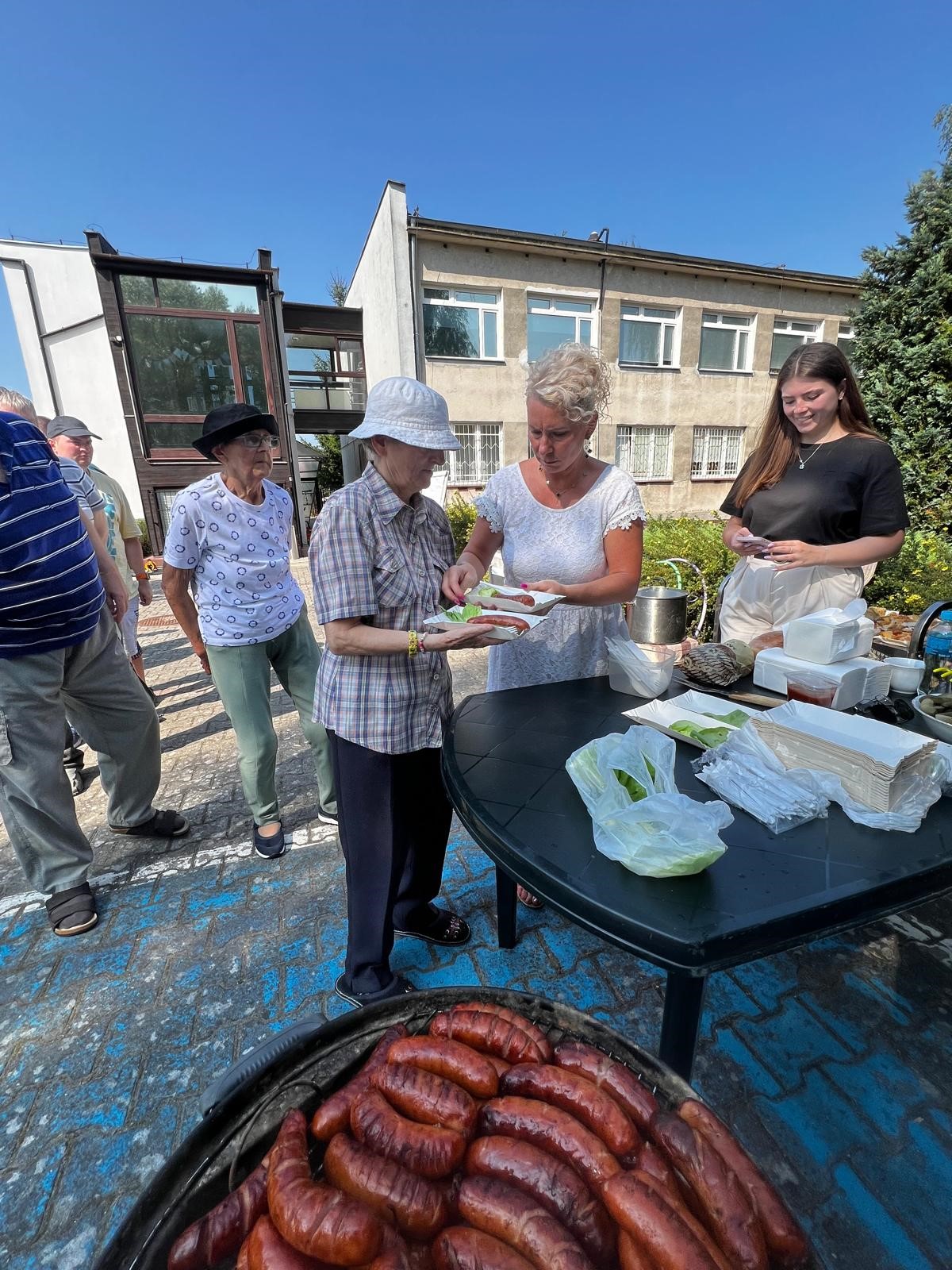 Na dole zdjęcia grill obłożony pieczonymi kiełbaskami. Na drugim planie kobieta w białej sukience wydaje upieczone kiełbaski na papierowych tackach osobom stojącym w kolejce