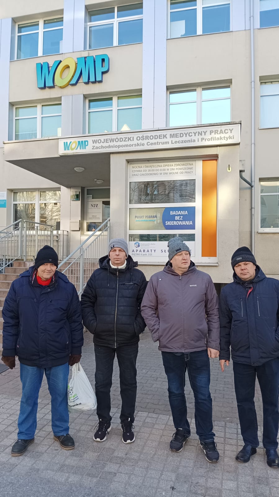 Czterech mężczyzn w kurtkach i czapkach stoi przed Wojewódzkim Ośrodkiem Medycyny Pracy.