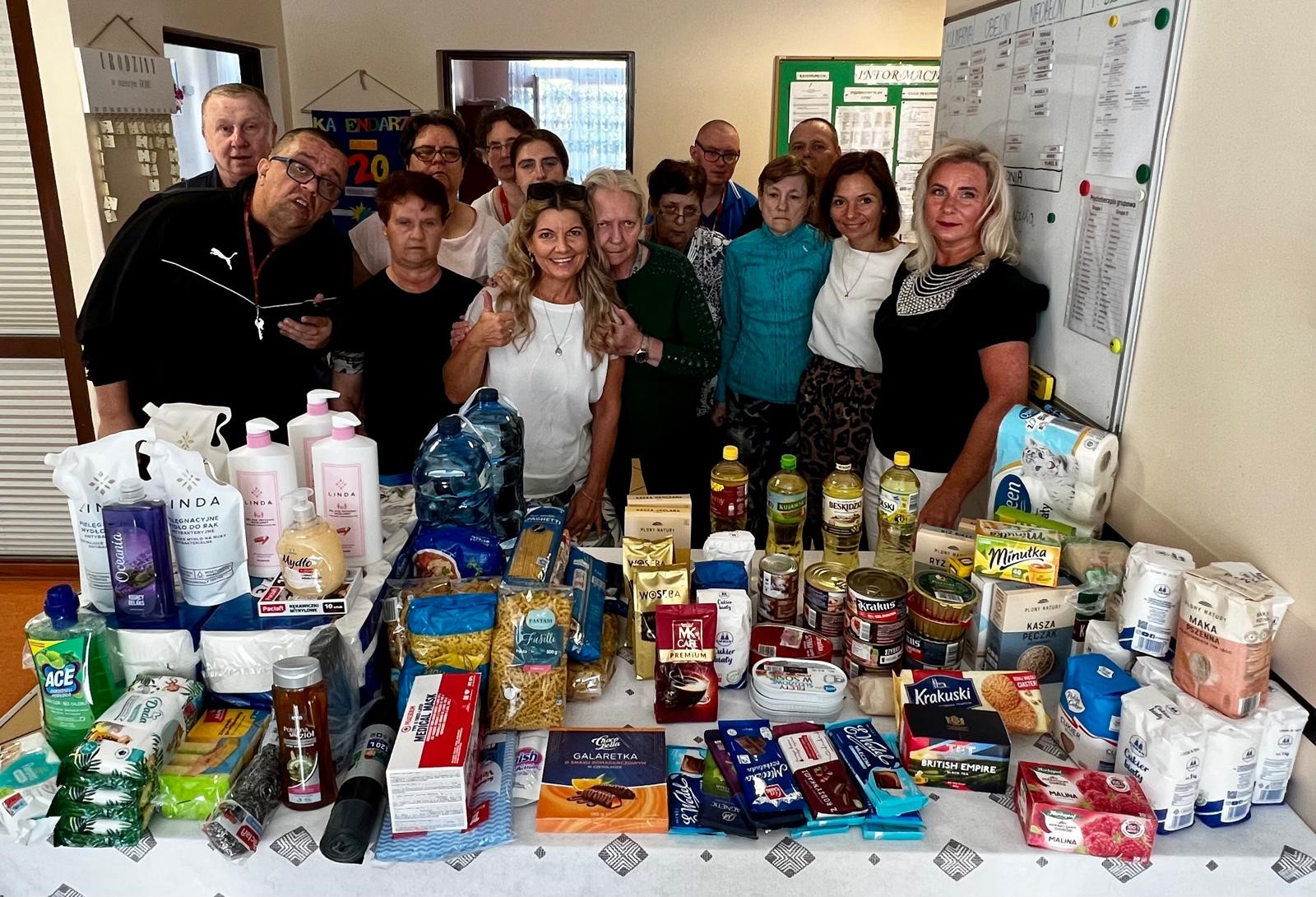 Na pierwszym planie stół z wieloma produktami spożywczymi i przemysłowymi takimi jak oleje, makarony, mąki, konserwy oraz mydła w płynnie. Na drugim planie grupa osób pozująca do zdjęcia. 