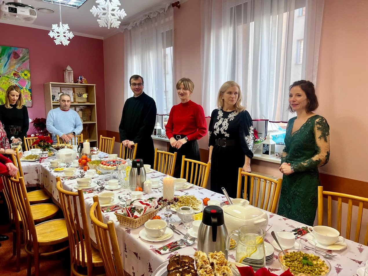 Grupa osób (kobiety i mężczyźni) ubrana w eleganckie stroje siedzi w dwóch salach, przy stołach zastawionych świątecznymi potrawami. 