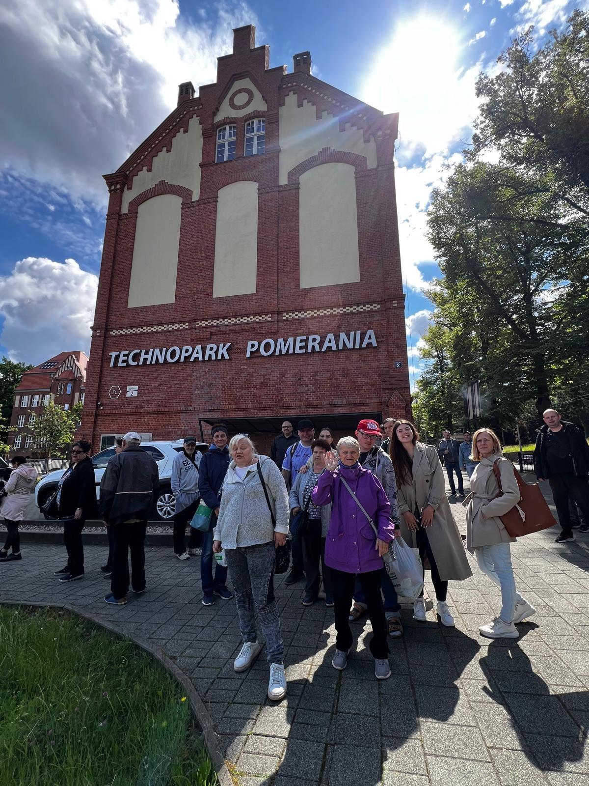 Grupa osób stoi przed ceglanym budynkiem z napisem Technopark. Jest słoneczna pogoda z umiarkowanym zachmurzeniem. 