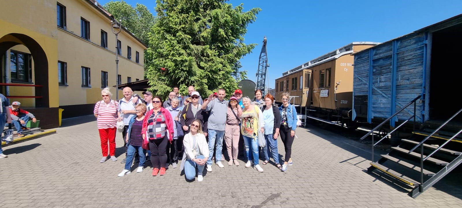 : Grupa kilkunastu osób pozuje do zdjęcia. Jest słoneczny, ciepły dzień. Po lewej stronie żółty budynek, po prawej stronie starodawne dwa wagony.