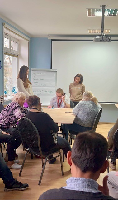 Na drugim planie siedzi mężczyzna z telefonem przy uchu. Po bokach stoją dwie kobiety, między nimi flipchart. Na pierwszym planie osoby siedzą tyłem do obiektywu i przyglądają się odgrywanej scence. 