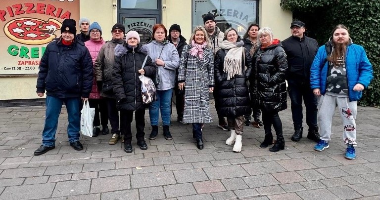 Na pierwszym planie grupa osób pozuje do zdjęcia. W tle budynek pizzerii.