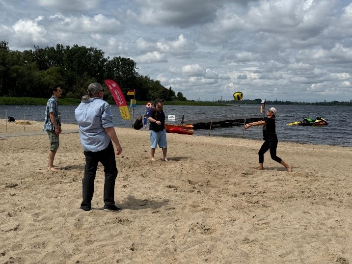 Cztery osoby na plaży grają w siatkówkę plażową. W tle jezioro z pomostem, drzewa i pochmurne niebo.