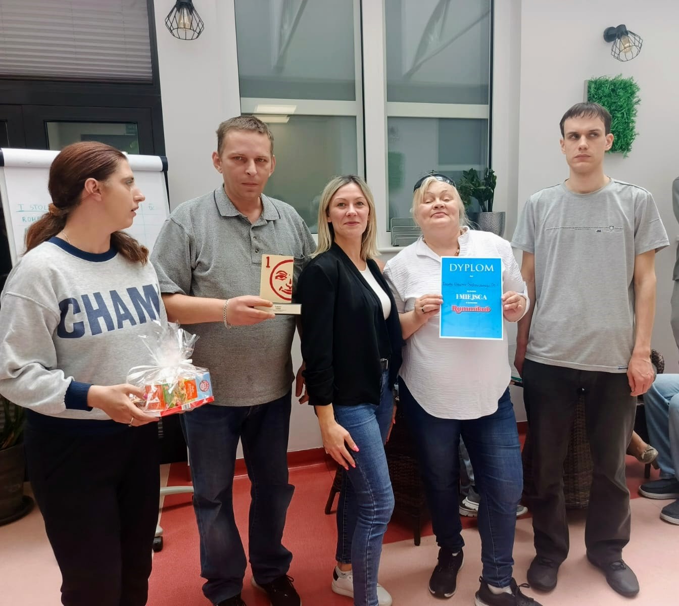 W jasnym pomieszczeniu pięć osób stoi i pozuje do zdjęcia. Jedna z nich trzyma niebieski dyplom w ręku.