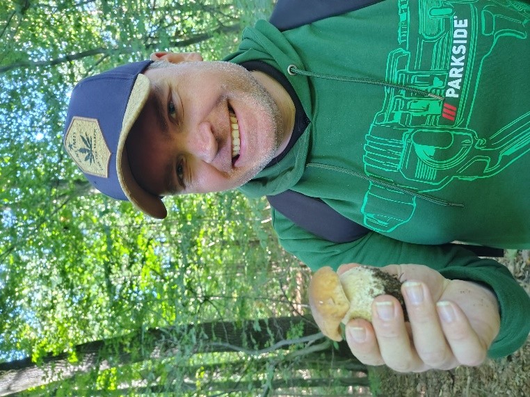 Uśmiechnięty mężczyzna w czapeczce i zielonej bluzie stoi w lesie i trzyma w ręku prawdziwka. 