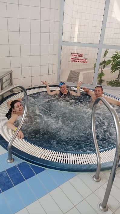 Trzy osoby relaksują się w jacuzzi w pomieszczeniu basenowym. Woda pieni się od bąbelków, a uczestnicy uśmiechają się i wyglądają na zadowolonych.