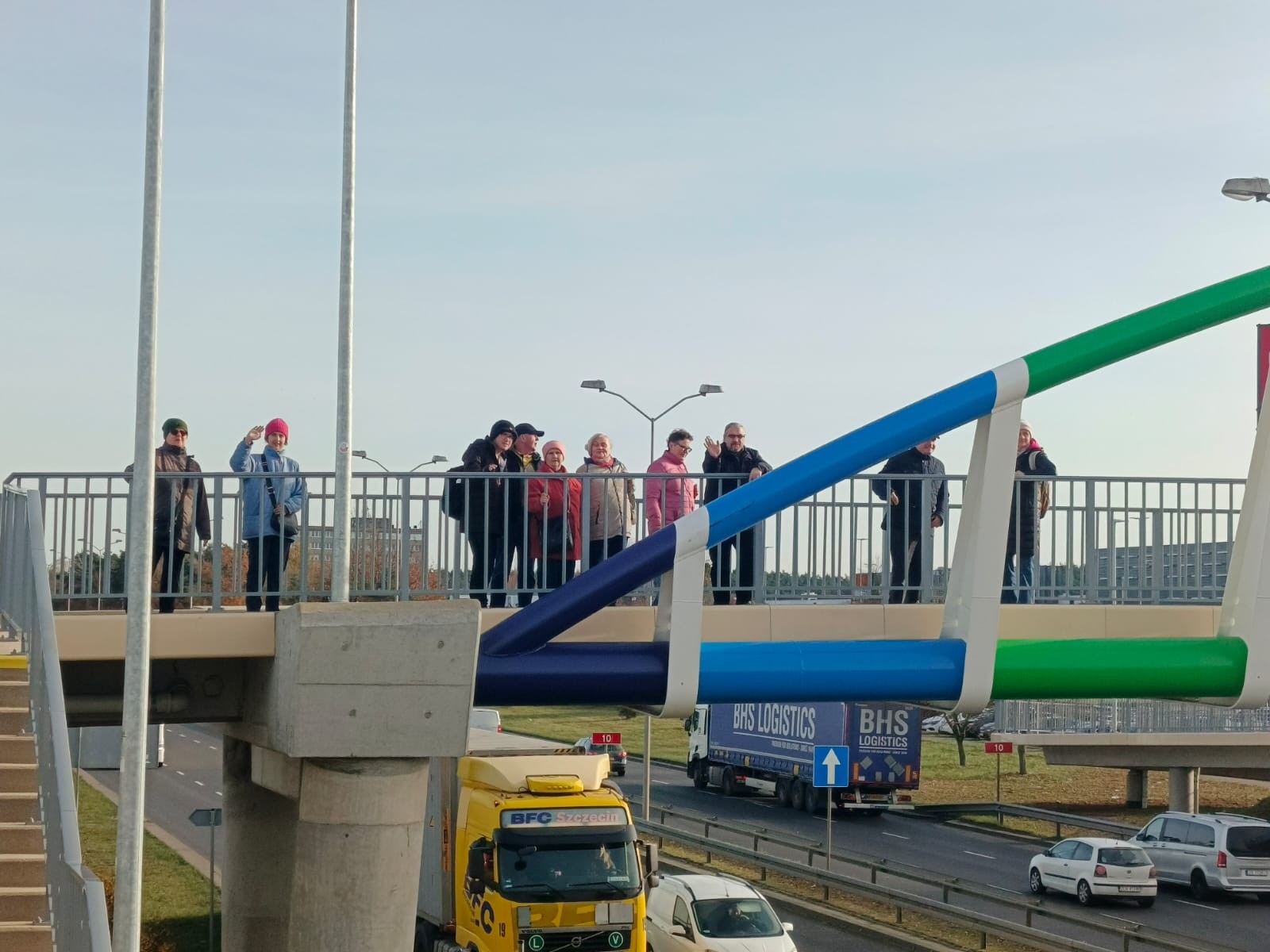 Grupa osób stoi na kolorowej kładce nad ruchliwą ulicą, pod spodem widoczne samochody i ciężarówki.
