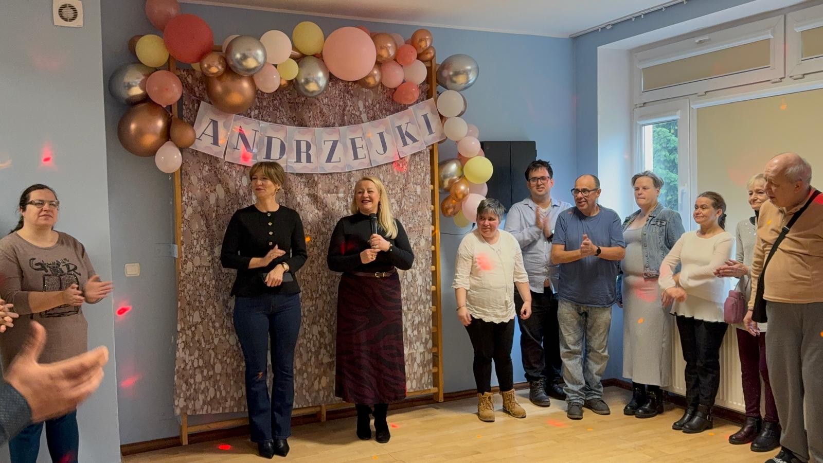Grupa osób bawi się w sali z niebieskimi ścianami, dekoracją z balonów i napisem „Andrzejki” w tle.