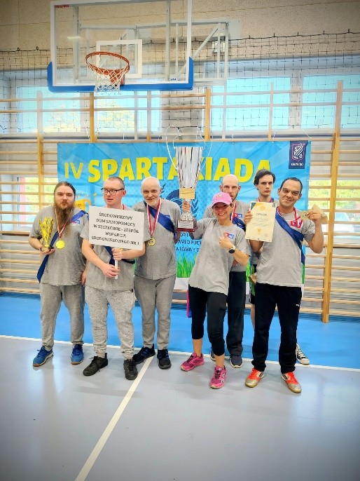 Grupa uśmiechniętych osób w szarych podkoszulkach stoi na sali gimnastycznej. Po środku kobieta wysoko trzyma duży puchar. 