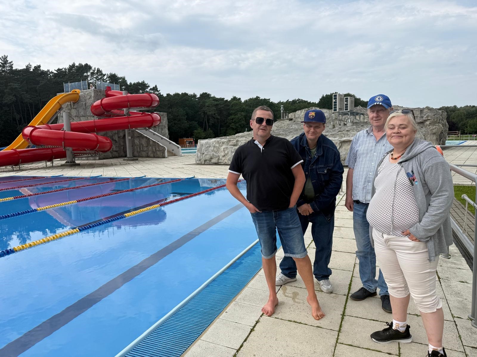 Po lewej stronie zdjęcia sportowy basen w plenerze, nad nim żółta i czerwona zjeżdżalnia. Po prawej stronie zdjęcia pozują 4 osoby.