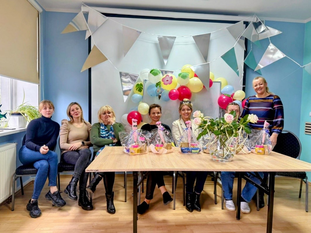 Grupa osób siedzi przy prostokątnym stole. Na stole znajdują się koszyki z upominkami, wazon z kwiatami. W tle widoczne są kolorowe balony i papierowe girlandy w kształcie trójkątów.