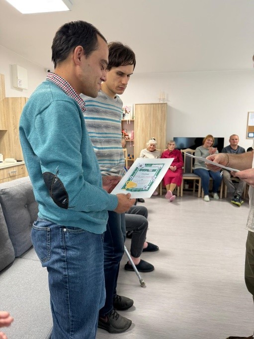 Dwie osoby stoją w jasnym pomieszczeniu, trzymając kolorowy dyplom. W tle widać kilka osób siedzących na krzesłach, szafki z dekoracjami oraz telewizor. 