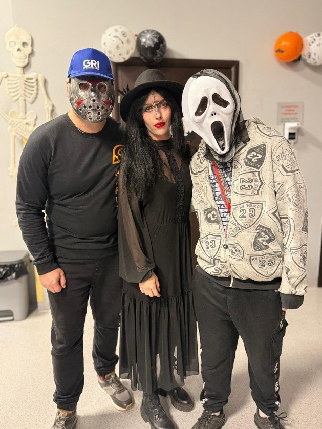 Trzy osoby stoją razem na tle halloweenowych dekoracji – widać papierowy szkielet na ścianie i balony w kolorach czarnym, białym i pomarańczowym. Stylizacje nawiązują do klimatu imprezy halloweenowej.