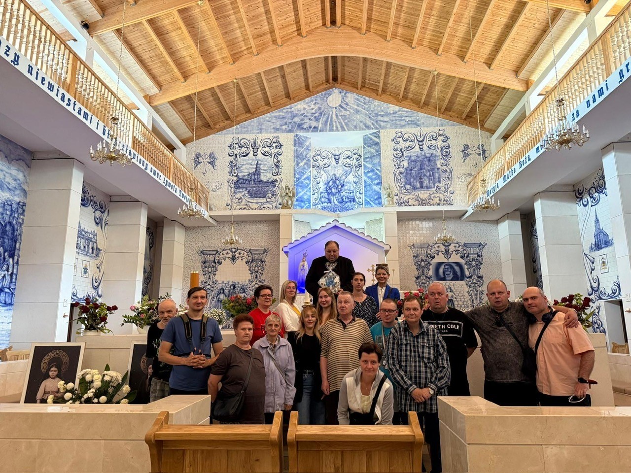 : Na zdjęciu widoczna jest grupa osób. W centralnym miejscu zdjęcia stoi ksiądz. W tle ołtarz o jasnej kolorystce oraz widoczna tylna	` ściana wewnętrzna Kościoła z białymi kaflami ceramicznym, które zdobione są ręcznymi malowidłami w kolorze niebieskim w stylu portugalskim.  