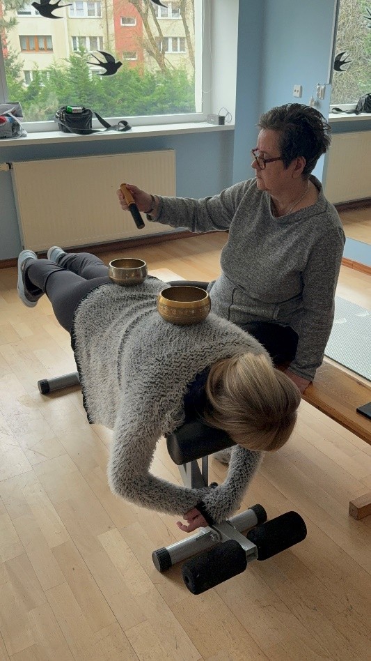 Osoba leży na ławce do ćwiczeń, na plecach stoją dwie misy tybetańskie, obok druga osoba uderza pałką w jedną z mis