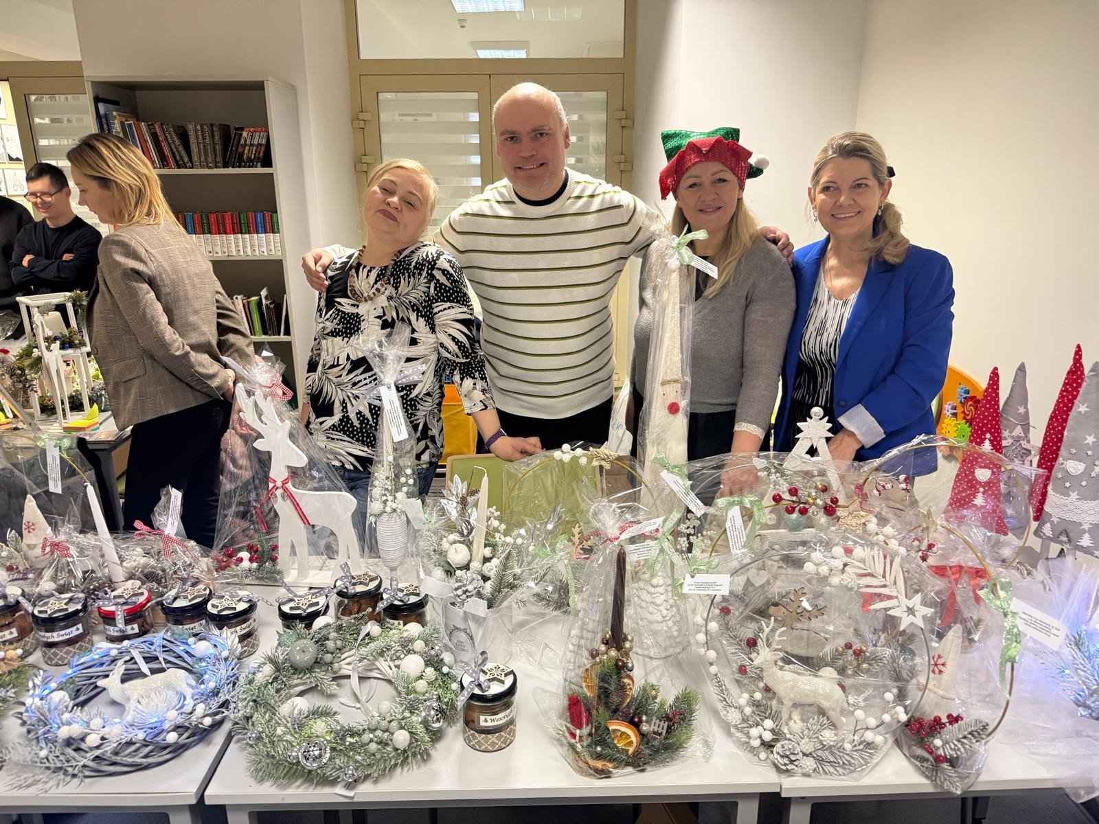 Na blatach znajdują się ozdobne wieńce, stroiki z gałązkami igliwia, choinki z różnych materiałów, słoiczki z piernikami oraz ręcznie wykonane dekoracje w kolorach bieli, srebra, czerwieni i zieleni. W tle widać osoby stojące przy stołach, a całość utrzymana jest w ciepłej, świątecznej atmosferze.