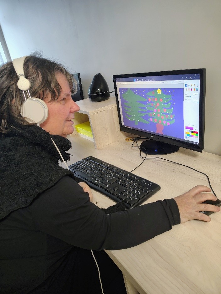 Osoba siedzi przy biurku, używając komputera z klawiaturą i myszką. Na monitorze widać program graficzny z rysunkiem dwóch choinek na niebieskim tle, ozdobionych bombkami i gwiazdą.