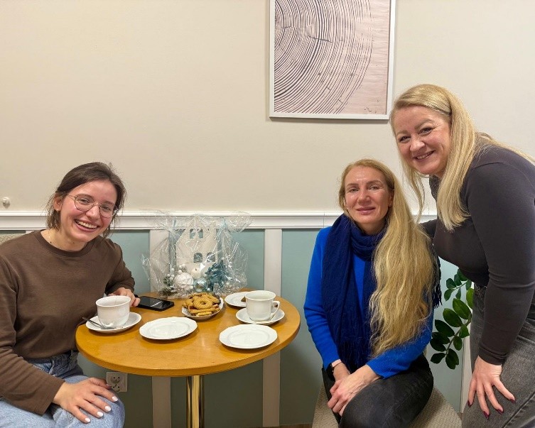 Trzy kobiety przy małym stoliku z filiżankami kawy i talerzykiem ciastek, obok świąteczna dekoracja owinięta w celofan.