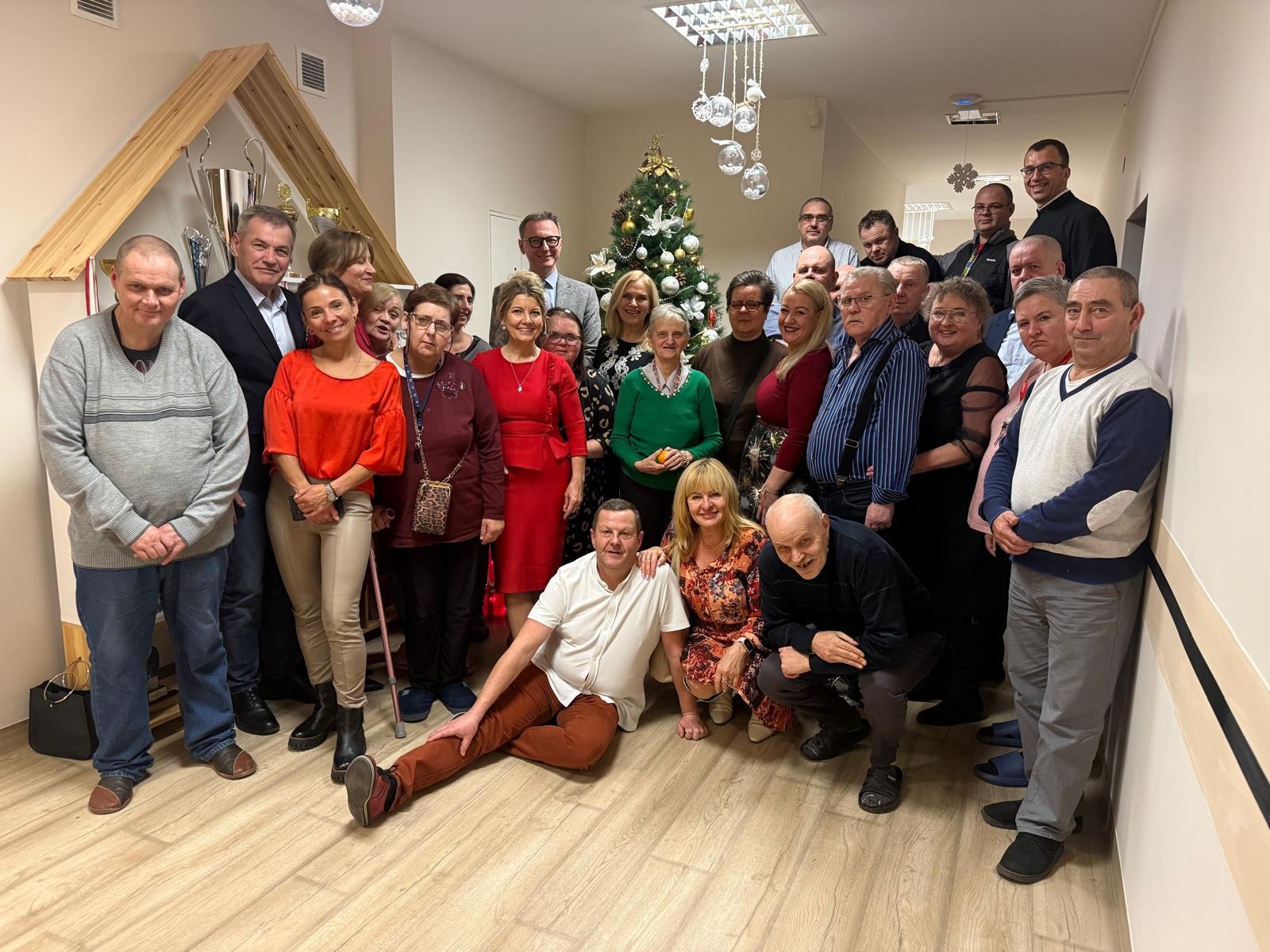 Grupa osób stoi i siedzi w jasnym pomieszczeniu, pozując do wspólnego zdjęcia przy udekorowanej choince z białymi i złotymi ozdobami. Nad nimi wiszą srebrne bombki, a po lewej stronie znajduje się drewniana konstrukcja z trofeami. Całość ma świąteczny, elegancki charakter.