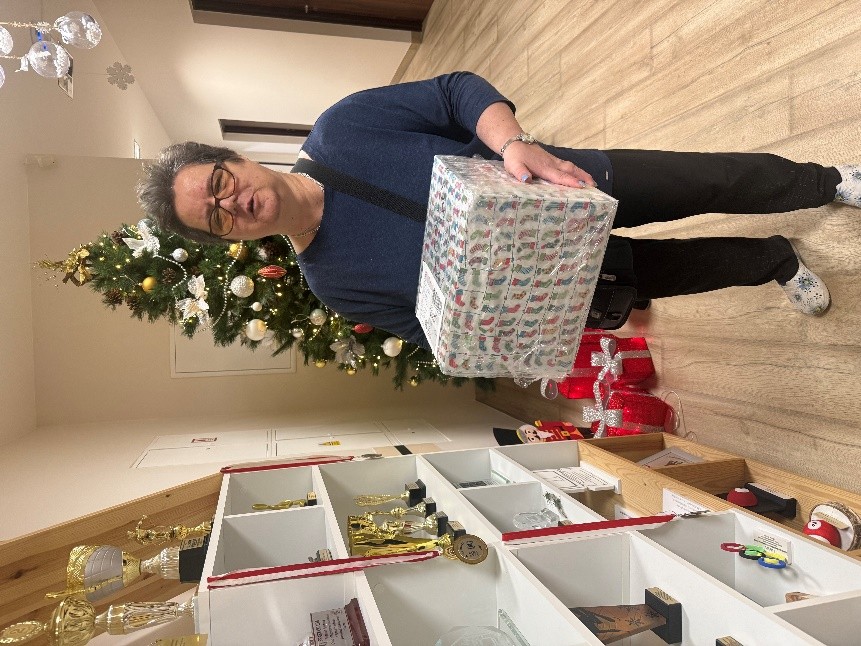 Osoba stoi obok półki z pucharami, trzyma duże, prostokątne pudełko owinięte w świąteczny papier. W tle widać choinkę ozdobioną bombkami i światełkami.