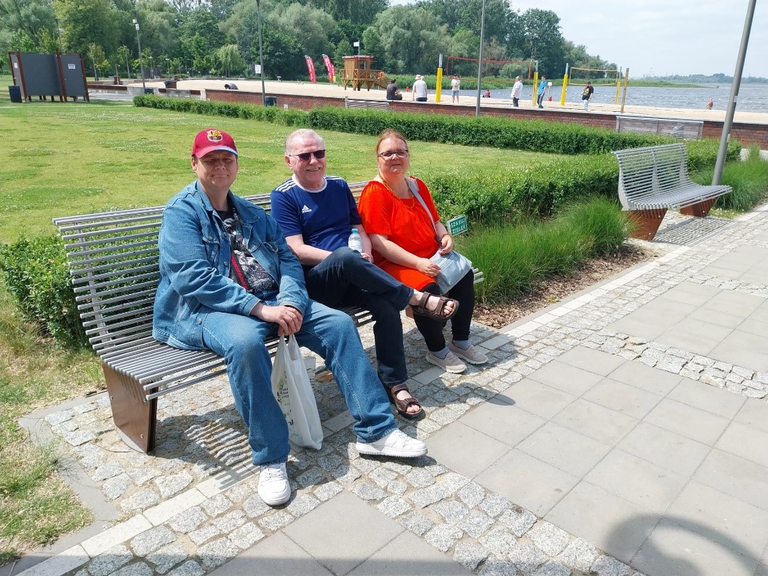 Na ławce przy chodniku siedzi dwóch mężczyzn i kobieta. Uśmiechają się patrząc w obiektyw. W tle zielona trawa i jezioro. 