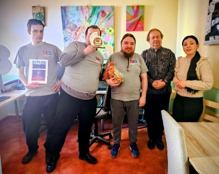 Grupa osób stoi w pomieszczeniu biurowym; jedna osoba trzyma dyplom, inna puchar, a kolejna upominek. W tle widoczne kolorowe obrazy oraz stanowiska komputerowe.