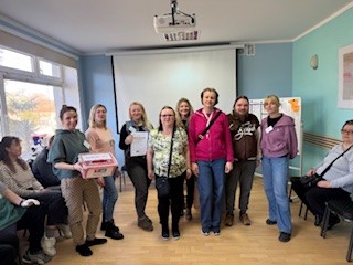 W sali z stoi grupa osób w pierwszym rzędzie, kobieta po lewej trzyma pudełko- urnę z kartami do głosowania. 