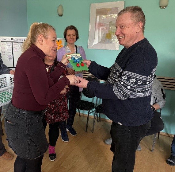 Na pierwszym planie stoi kobieta i przekazuje uśmiechniętemu mężczyźnie kolorową kartkę ozdobną, a reszta stoi obok i obserwuje.