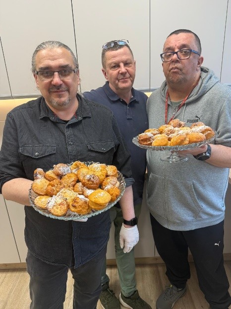 Trzech mężczyzn stoi w kuchni, a dwie z nich trzymają duże szklane patery wypełnione małymi, złocistymi pączkami posypanymi cukrem pudrem.