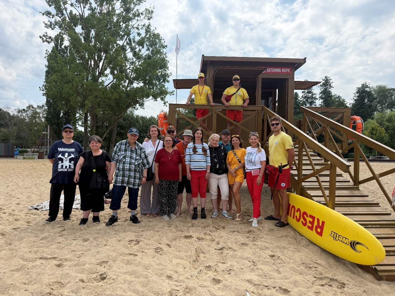 Na piaszczystej plaży stoi grupa uczestników. Za nimi znajduje się drewniana budka ratowników WOPR. W tle widoczne są zielone drzewa oraz pochmurne, zachmurzone niebo. Uczestnicy znajdują się na otwartej przestrzeni nad wodą.