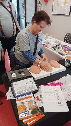 Na pierwszym planie biurko z ulotkami informacyjnymi oraz trzy fantomowe piersi. Za biurkiem stoi kobieta ćwicząca samobadanie piersi na fantomie