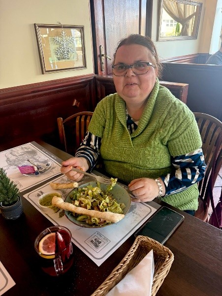 Uśmiechnięta kobieta siedzi przy stoliku w restauracji, je sałatkę z grzankami; na stole herbata w szklance.