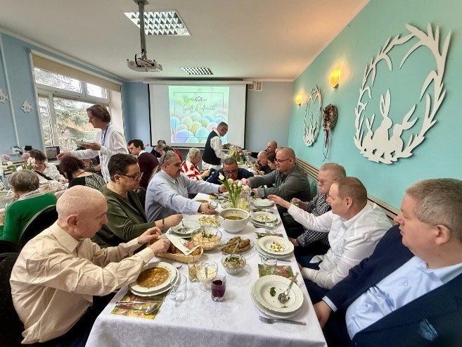 Grupa osób siedzi przy długim, nakrytym białym obrusem stole i wspólnie je wielkanocny posiłek w jasnej, udekorowanej sali. Na stole stoją zupy, jajka i sałatki. W tle widać ekran z wielkanocną grafiką.